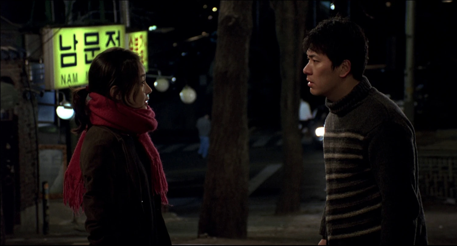 A man and a woman stand outside at night, facing each other. Trees and neon signs are visible in the background.